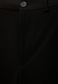 Gros plan sur un pantalon en tissu noir avec un bouton noir à quatre trous et des coutures visibles au niveau de la ceinture et de la fermeture éclair.