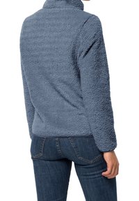 Polarowy pulower w miękkim, fakturowanym niebieskim kolorze z wysokim kołnierzem i długimi rękawami. Noszony na ciemnych dżinsach, prezentuje luźny krój.