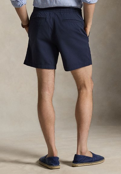 Μπλε σκούρο σορτς με ελαστική μέση, υφασμάτινη υφή και διπλές πίσω τσέπες, συνδυασμένα με μπλε σκούρα παπούτσια χωρίς κορδόνια.