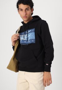 Hoodie en coton noir avec un design graphique bleu portant l'inscription "Tommy Jeans". Dispose d'une poche à l'avant et de poignets côtelés. Modèle tenant un sac beige.