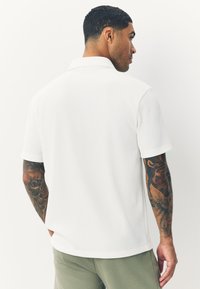 White ribbed short-sleeve polo with a structured collar and subtle texture, paired with olive green pants. Notable tattoo design on sleeve.