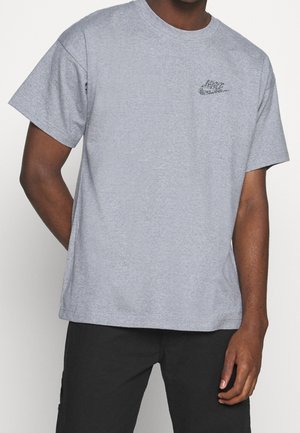 Mann trägt ein hellgraues Nike-T-Shirt mit schwarzen Hosen und steht mit einer Hand hinter dem Rücken vor einem einfarbigen Hintergrund.