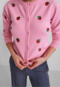 Kvinna som knäpper en ljusrosa knäppt cardigan med broderade röda jordgubbar, tillsammans med mörkgrå byxor, mot en enfärgad grå bakgrund.