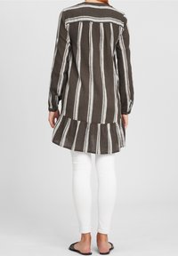 Brązowo-białe paskowane tunika z długimi rękawami, teksturowany materiał i falbana na dole, dopasowana do białych legginsów i czarnych sandałów.