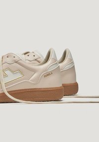 Baskets avec une tige en cuir beige clair, semelle en caoutchouc naturel, et coutures jaunes contrastantes avec le logo "BURELA" sur le talon.