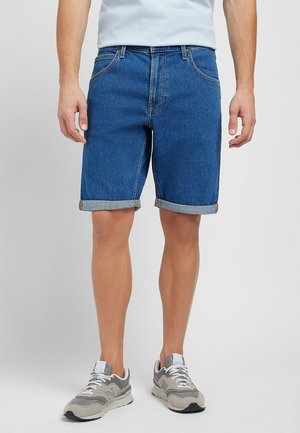 Mand iført op rullede blå denimshorts, grå og hvide sneakers og en lyseblå skjorte står mod en ensfarvet baggrund.