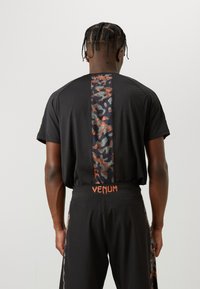 Črna športna majica s kamuflažnim vzorcem na sredini ter logotipom "VENUM" v oranžni barvi na hrbtnem delu in pasu usklajenih kratkih hlač.