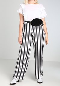 Kvinde iført hvid top med flæser, sort og hvidt vertikalt stribet vide bukser, hvide sko og sort oval bæltetask.