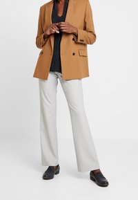 Blazer doppiopetto marrone, top nero a scollo a V, pantaloni svasati grigio chiaro e mocassini neri, che mostrano un design su misura e texture morbide.