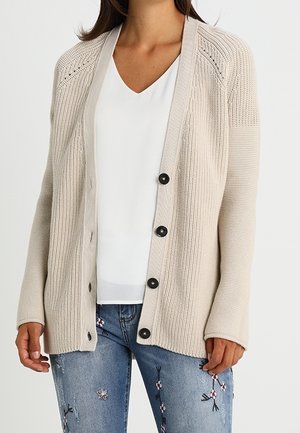Femme portant un cardigan en maille beige avec des boutons noirs sur un haut blanc à col en V et un jean bleu avec broderies florales.