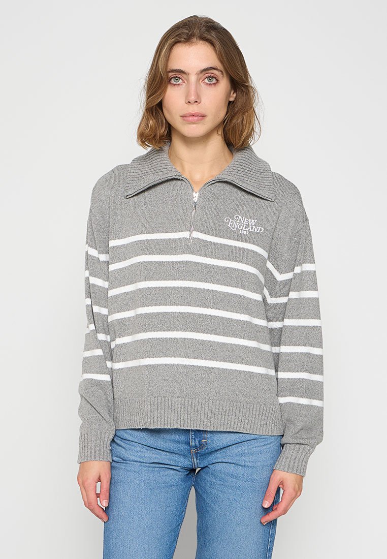 Pull en tricot gris avec des rayures blanches horizontales, un col haut et une fermeture éclair à l'avant. Présente un texte brodé sur la poitrine.