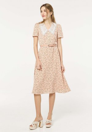 Woman wearing a pink floral midi dress with puff sleeves, white lace collar, a matching belt, and white high-heeled braided sandals.