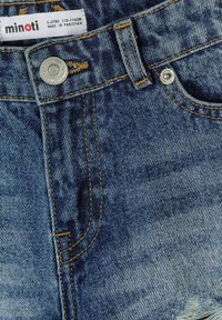 Jean en denim bleu avec boutons en métal, coutures jaunes, poche avant et étiquette indiquant "minoti" 5-6 ans, fabriqué au Pakistan.