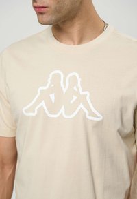 Béžové tričko z bavlny s veľkým bielym siluetovým grafickým motívom dvoch sediacich postáv. Okrúhly výstrih a krátke rukávy.