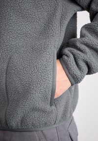 Grå fleecetröja med texturerat tyg, sidoficka och kontrasterande kant längs sömmar och ärmslut. Närbild med fokus på ärmen och fickan.