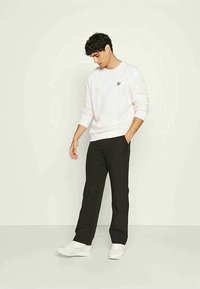 Lichtroze sweatshirt met een klein geel logo, zwarte broek en witte sneakers. Model staat met handen in de zakken, casual houding.