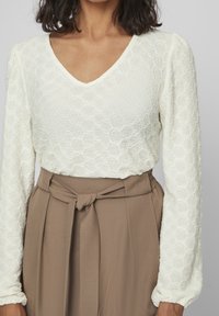 Haut blanc texturé à manches longues avec un col en V, présentant un motif en nid d'abeille. Assorti à un pantalon marron taille haute et une ceinture nouée.