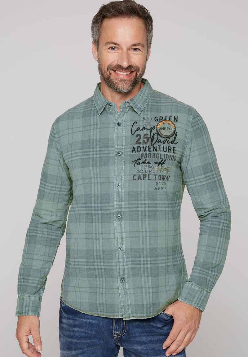 Camisa de mangas largas a cuadros verdes con botones en el frente, con gráficos de texto en el pecho. Tejido suave con dobladillo curvado y puños abotonados.