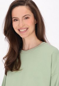 Top verde chiaro a girocollo realizzato in un tessuto morbido, con una vestibilità rilassata e spalle leggermente scese, caratterizzato da una texture liscia e un design semplice.