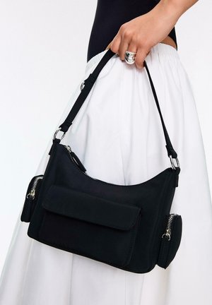 Main tenant un sac d'épaule noir avec une poche à rabat à l'avant et deux poches latérales zippées, contre une jupe blanche et un haut noir.