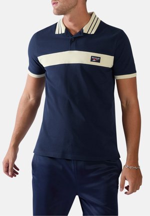 Mann trägt ein dunkelblaues Reebok-Poloshirt mit cremefarbenem Streifen über der Brust sowie am Kragen und an den Ärmelsäumen, steht mit entspannten Armen.
