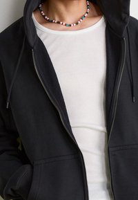 Svart zip-up hoodie med en mjuk textur och två framfickor, lager över en vit ribbad långärmad tröja. Färgglad pärlhalsband som accent.