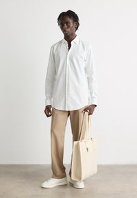 Hvid skjorte med knapper, beige bukser og hvide sneakers; holder en lysebeige totebag med et struktureret design. Glat stof og rene linjer.