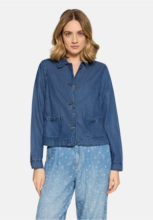 Młoda kobieta o blond włosach do ramion, ubrana w niebieską kurtkę jeansową zapinaną na guziki oraz jasne, niebieskie spodnie w paisley, stojąca na białym tle.