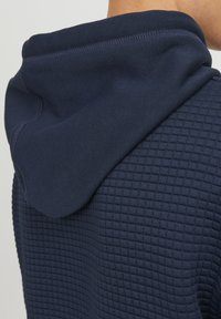 Dunkelblaue Kapuzenjacke mit einem strukturierten, gitterartigen Muster. Verfügt über eine weiche, verstellbare Kapuze und glatte Nähte entlang der Nähte.