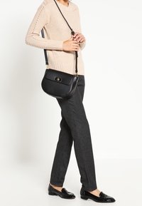 Frau trägt einen beigen Strickpullover, dunkle graue Hose, schwarze Slipper und eine schwarze Umhängetasche mit einem goldenen Verschluss.