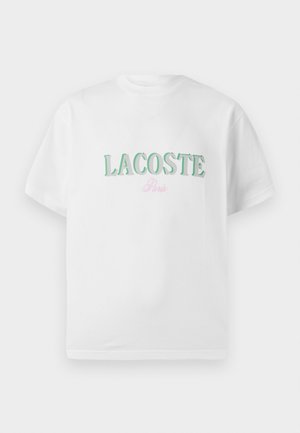 Bijela pamučna majica s zelenim natpisom "LACOSTE" i ružičastim "Pariz" u zakrivljenom dizajnu. Kratki rukavi i okrugli izrez. Glatka tekstura.