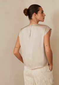 Beżowy top bez rękawów o delikatnym połysku, z małym otworem w kształcie dziurki z tyłu. Materiał ma lekką strukturę, w zestawieniu z wzorzystymi spodniami.