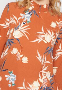 Abito color ruggine con colletto arricciato. Presenta un motivo floreale in bianco, blu e verde, con maniche lunghe e una texture morbida.