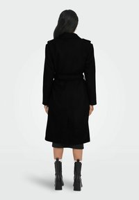 Manteau long en mélange de laine noire avec ceinture, revers structurés et fente arrière. Porté sur une jupe mi-longue et assorti à des bottes noires à talons blocs.