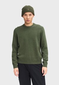 Maglione verde oliva con un classico scollo rotondo e polsini a costina, abbinato a un berretto a coste coordinato. Tessuto semplice e strutturato.