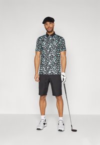 Kortermet svart blomster polo skjorte med teal og hvite mønstre, kombinert med svarte shorts og hvite treningssko. Holder en golfkølle.