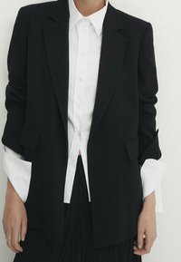 Zwarte op maat gemaakte blazer met een gestructureerd ontwerp, voorzien van twee voorzakken en opgerolde witte shirtmanchetten die onder een wit overhemd worden gedragen.