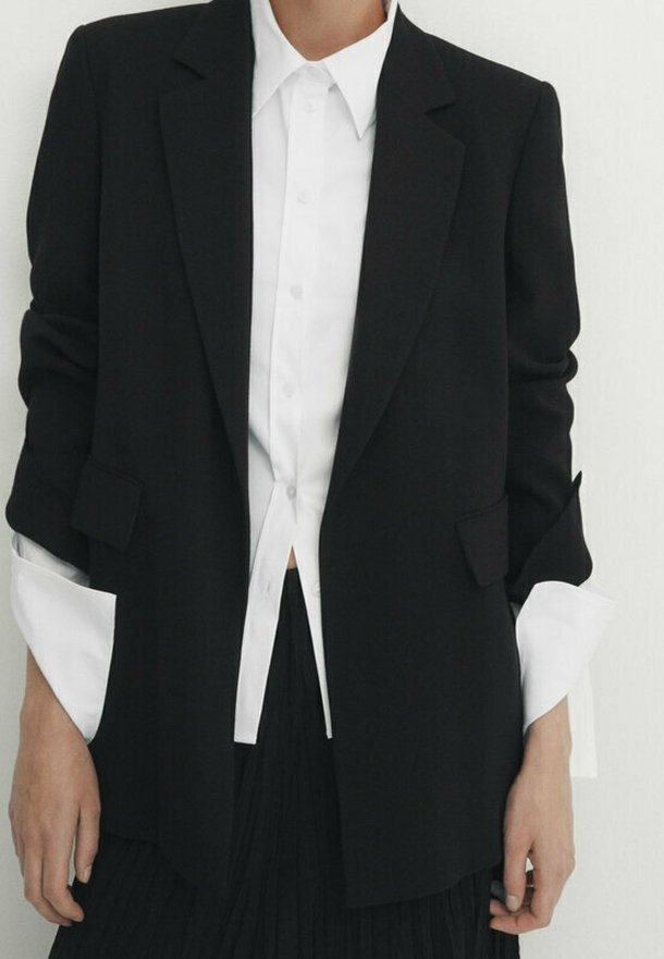 Zwarte op maat gemaakte blazer met een gestructureerd ontwerp, voorzien van twee voorzakken en opgerolde witte shirtmanchetten die onder een wit overhemd worden gedragen.