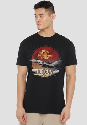 Junger Mann, der ein schwarzes Top Gun T-Shirt mit einem Kampfjet und dem Text "Ich spüre das Bedürfnis... das Bedürfnis nach Geschwindigkeit!" trägt, steht vor einem grauen Hintergrund.