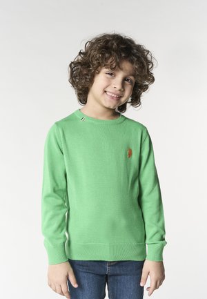 Bambino sorridente con capelli ricci che indossa un maglione verde a maniche lunghe e jeans blu, in piedi davanti a uno sfondo bianco semplice.