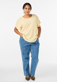 Keltainen, lyhythihainen toppi, valmistettu kevyestä kankaasta; korkeavyötäröiset siniset farkut, joissa kirjailtuja kukkaisia yksityiskohtia; ruskeat kengät.