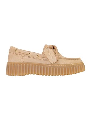 Mocassin en daim beige avec semelle épaisse en caoutchouc crantée, lacets noués en nœud papillon à l'avant et étiquette décorative attachée aux lacets.