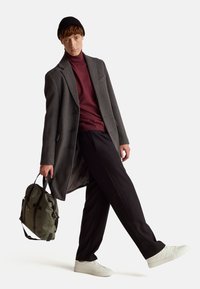 Cappotto grigio sopra un dolcevita bordeaux, pantaloni neri e sneakers bianche. Tiene una borsa verde con manici neri. Dettagli del tessuto testurizzati.