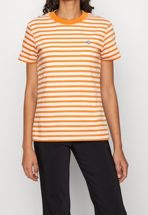 Osoba ubrana w biało-pomarańczową koszulkę w poziome paski na krótkim rękawie i czarne spodnie, stojąca na jednolitym tle.