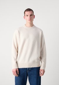 Kremfarget, teksturert genser med rund hals, ribbestrikkede mansjetter og fald. Kombinert med blå denim, med et lite logo-merke.