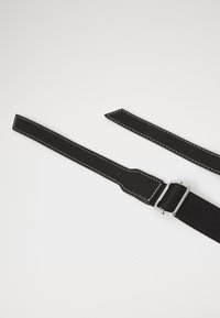 Czarny skórzany pasek z białymi szwami, prostokątną srebrną klamrą i regulowanymi końcami na białym tle.