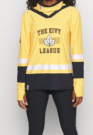 Person trägt ein gelb-schwarzes Langarmshirt mit der Aufschrift "THE EIVY LEAGUE" und einem Blattmotiv, kombiniert mit schwarzen Hosen.