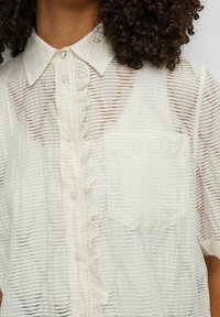 Lätt, genomskinlig krämfärgad blus med vertikala ränder, volangdetalj på framtill och korta ärmar. Har en enkel bröstficka.