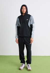 Young man wearing a black and grey Nike hoodie with the hood up and matching black Nike sweatpants and white sneakers, standing on green floor.