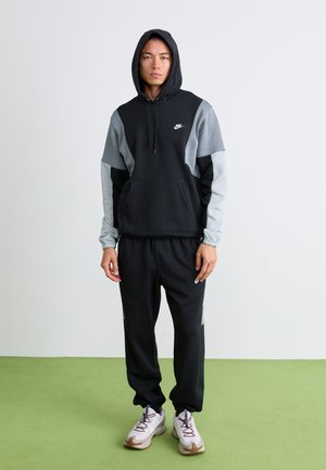 Jeune homme portant un sweat à capuche Nike noir et gris avec la capuche relevée, associé à un pantalon de survêtement Nike noir et des baskets blanches, se tenant sur un sol vert.